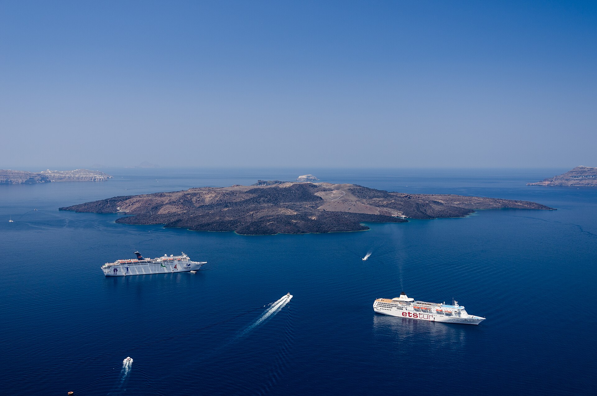 Cruise ships anchored in Santorini's blue caldera with the volcanic island of Nea Kameni beyond