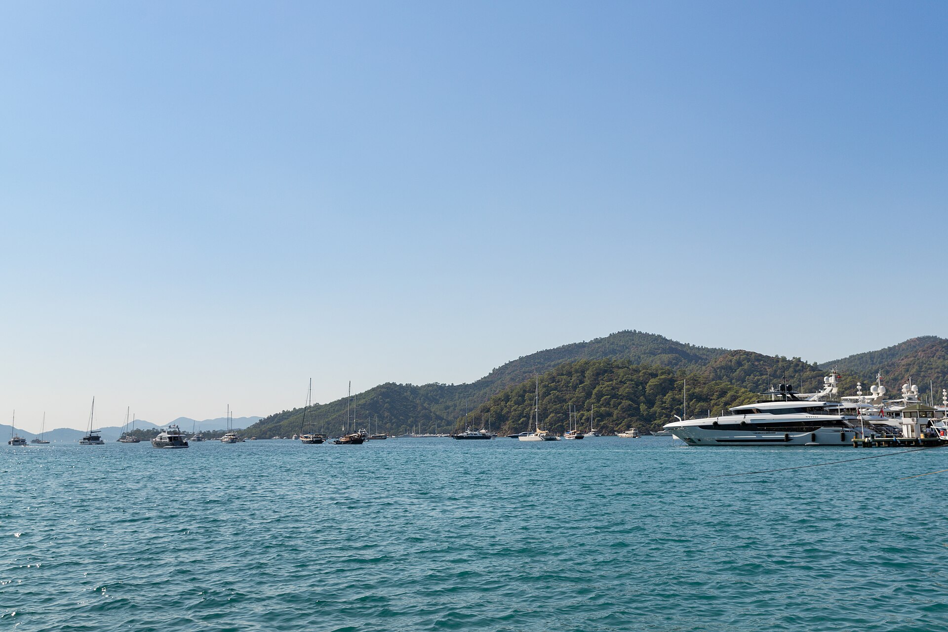 Sailing yachts and small passenger vessels across turquoise Göcek Bay beside a wooded island