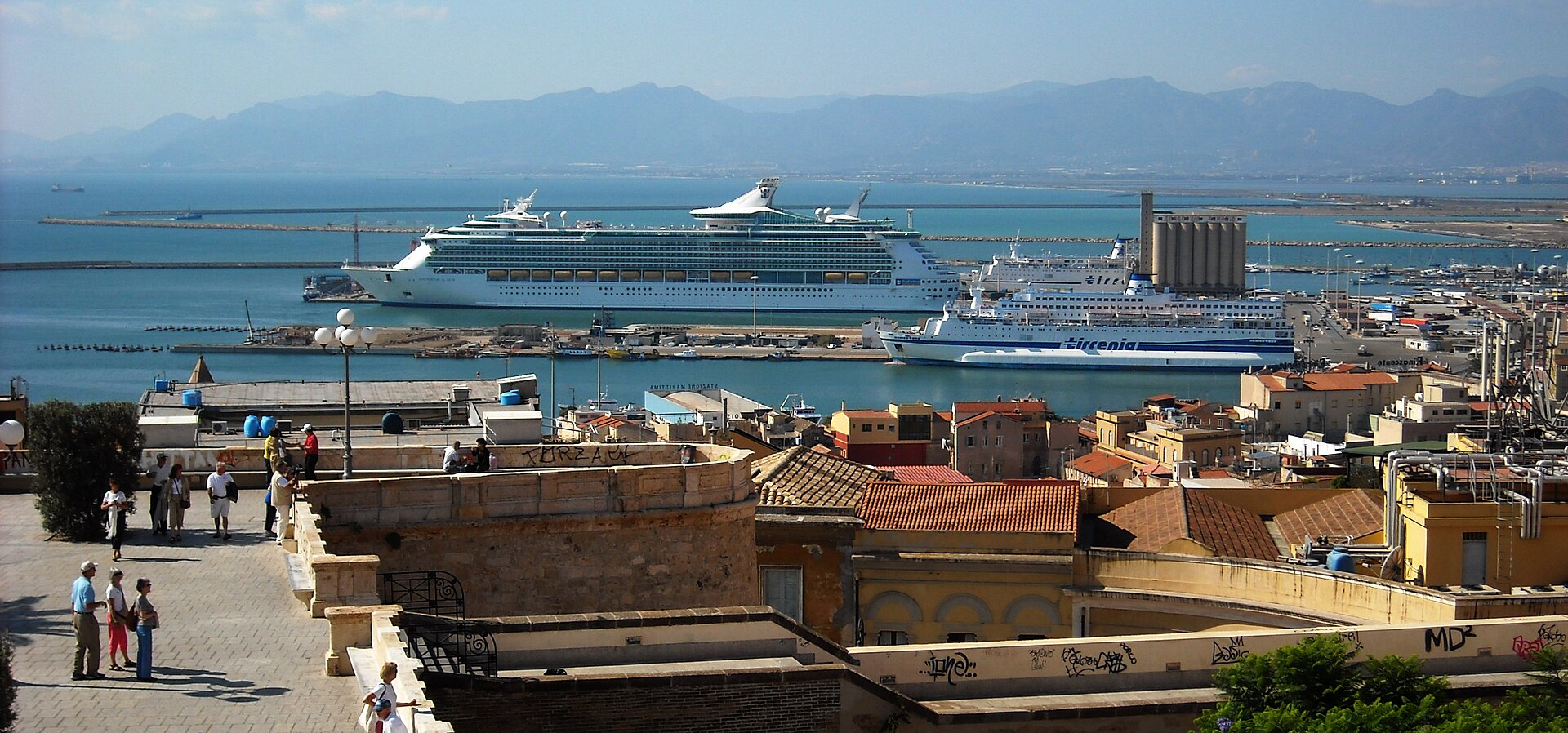 Cruise ship and passenger ferry in Cagliari harbor below city roofs, with the gulf and Sardinian mountains beyond
